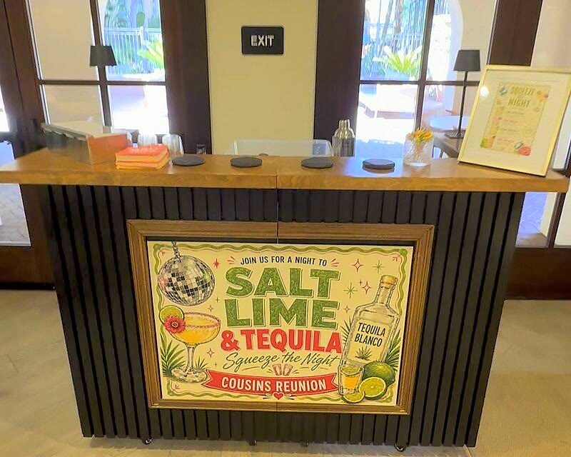 The Salt Rim's black slat-front mobile bar with a wood top, set up at a venue with a client's custom framed banner on the front