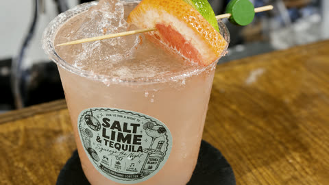 A finished cocktail over ice with a grapefruit wedge and lime on a salted rim, in a cup carrying the event's own sticker