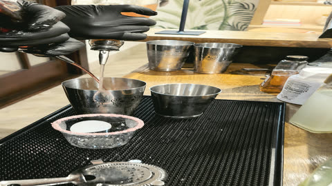 A bartender's gloved hands salting the rim of a glass over the bar mat, mixing tins and a strainer beside it