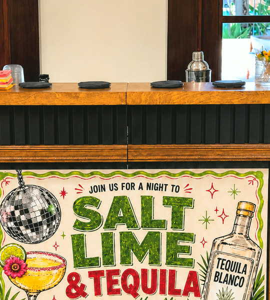The Salt Rim bar top set before doors open &mdash; bar mats, a cocktail shaker and stacked cups on the wood counter, the client&rsquo;s framed &lsquo;Salt Lime & Tequila&rsquo; banner on the black slat front below