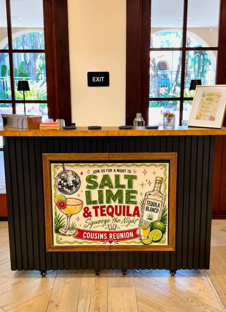 The Salt Rim mobile bar set up at a client's venue — black slat front, wood top, a custom framed banner reading 'Salt Lime & Tequila', and the client's printed menu on the bar, arched windows and greenery behind